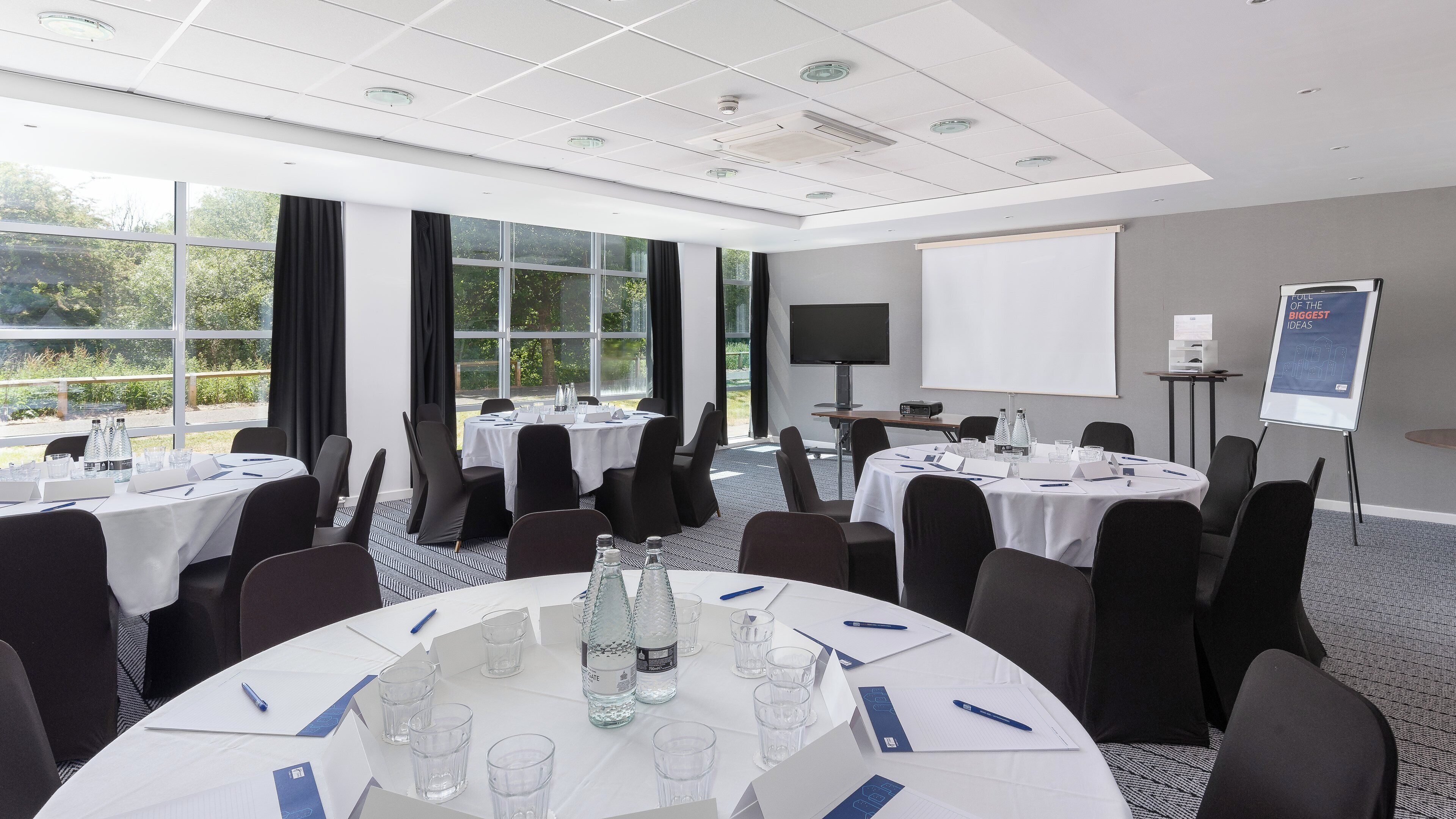 Un salón de conferencias con varias mesas, sillas y una pantalla para proyector.