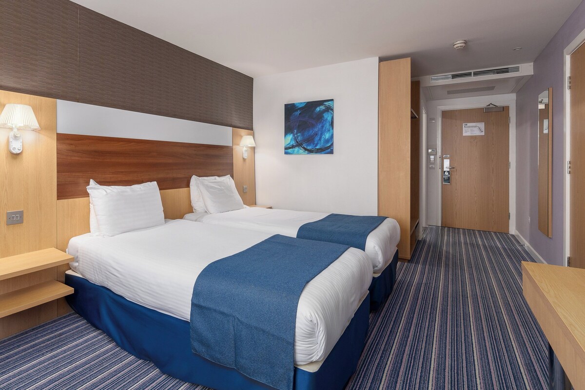 A hotel room with a large bed, two bedside tables, a nightstand, a wall-mounted lamp, a painting, and a door with a mirror.