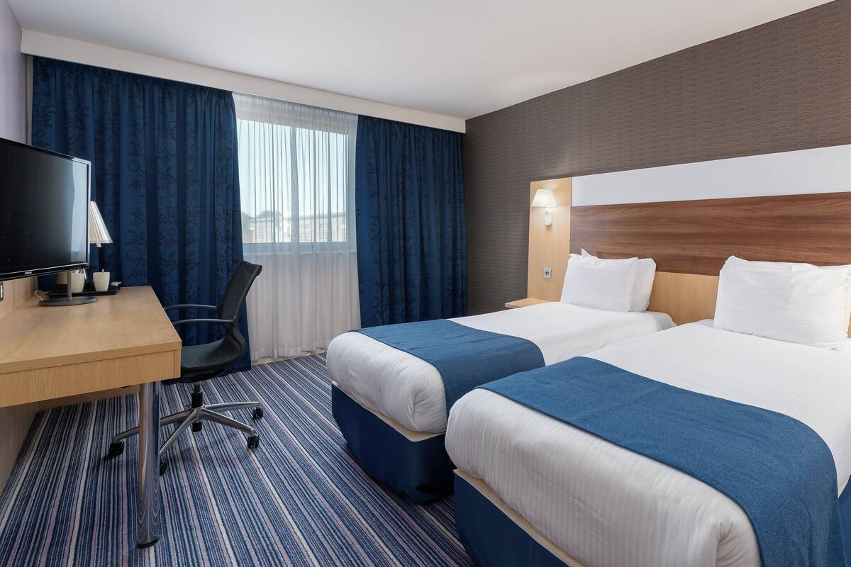 A hotel room with two beds, a desk, a chair, a TV, and a window with curtains.