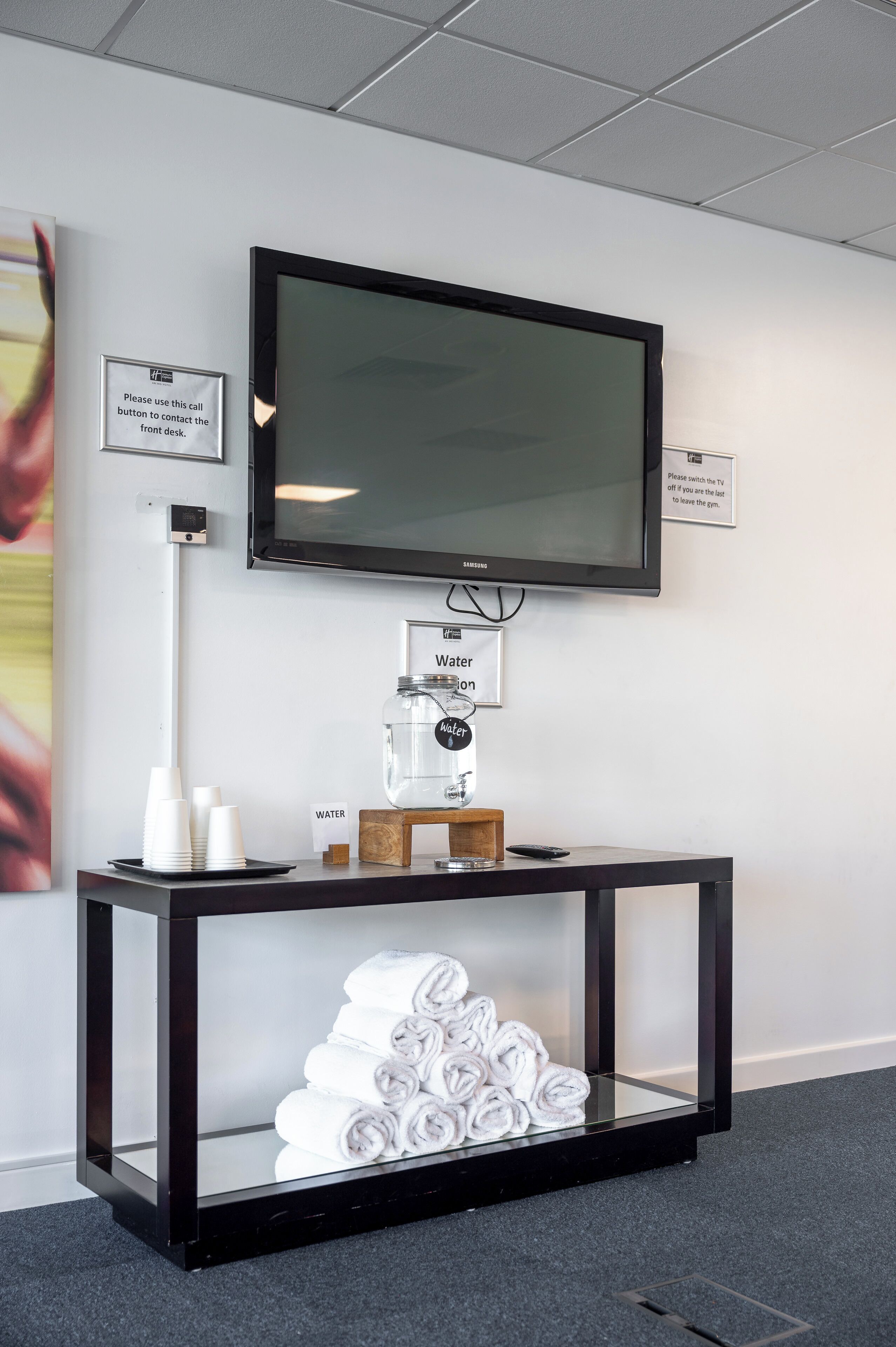 A modern reception area with a flat-screen TV mounted on the wall, a water dispenser, and a console table with folded towels.