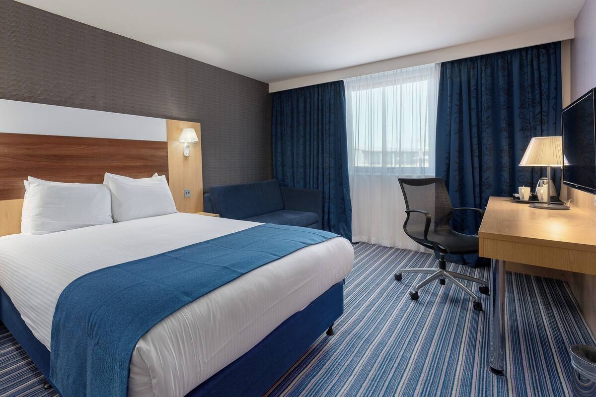 A hotel room with a large bed, a desk with a chair, a TV, and a window with curtains.