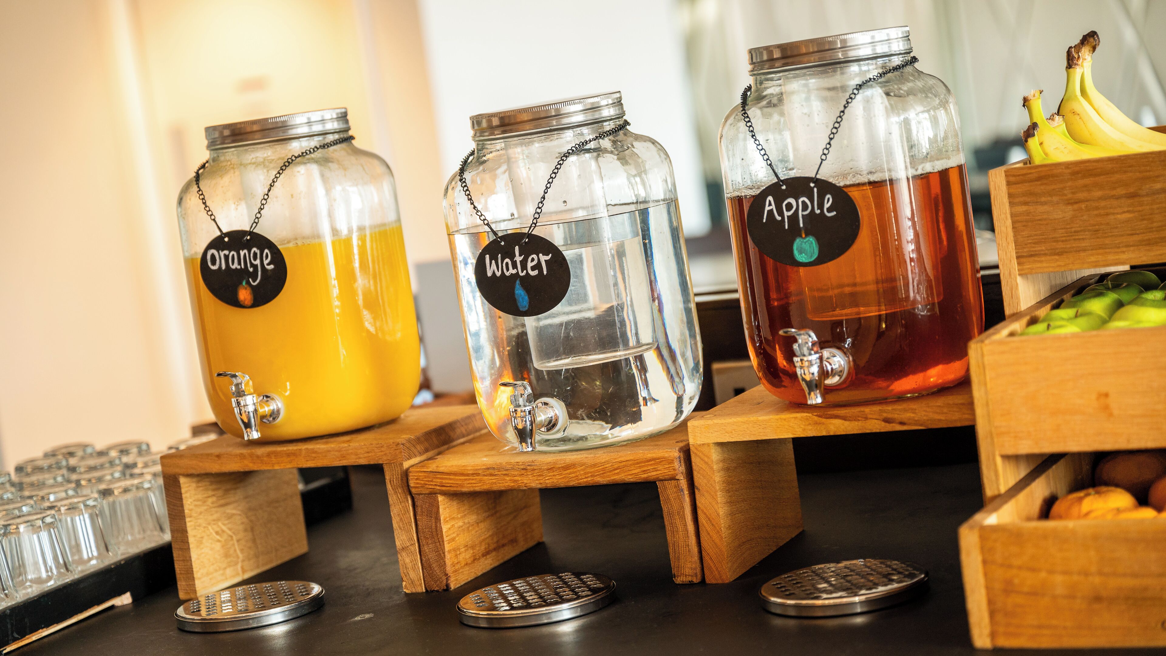 Three glass jars with taps on wooden stands, labeled orange, water, and apple.
