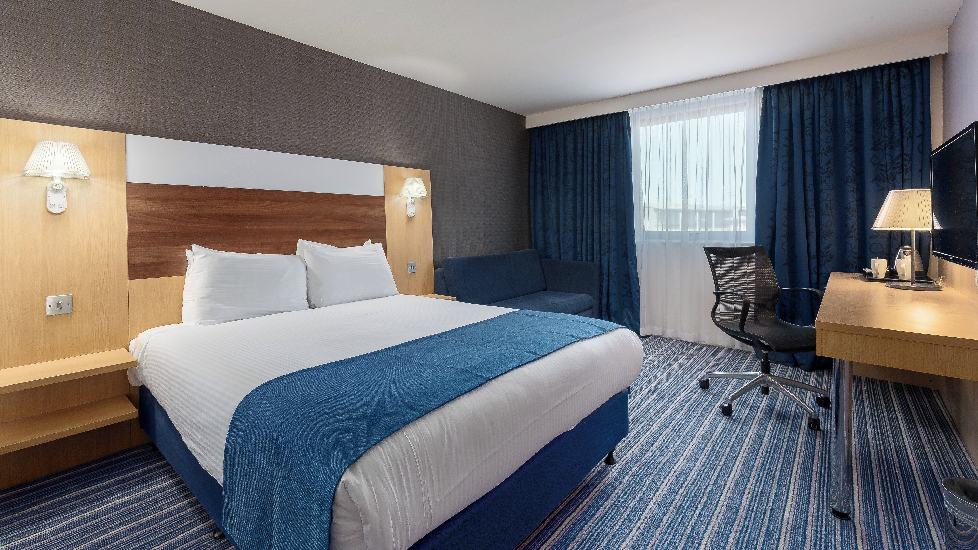 Een hotelkamer met een groot bed, een bureau met stoel, een televisie en een raam met gordijnen.