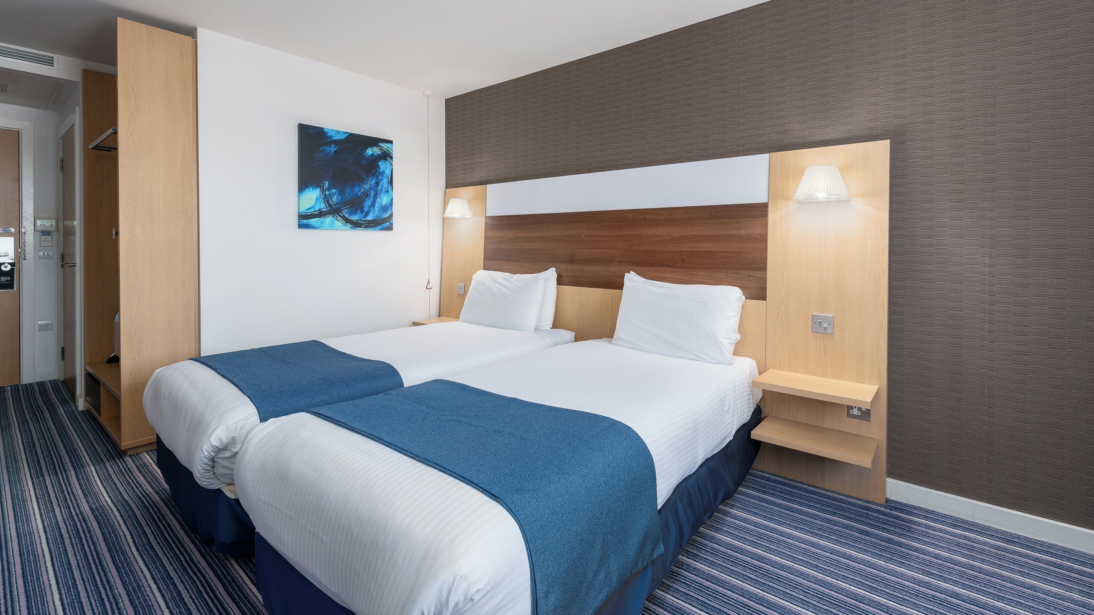 A hotel room with a double bed, a headboard, bedside tables, a wall-mounted artwork, and a carpeted floor.
