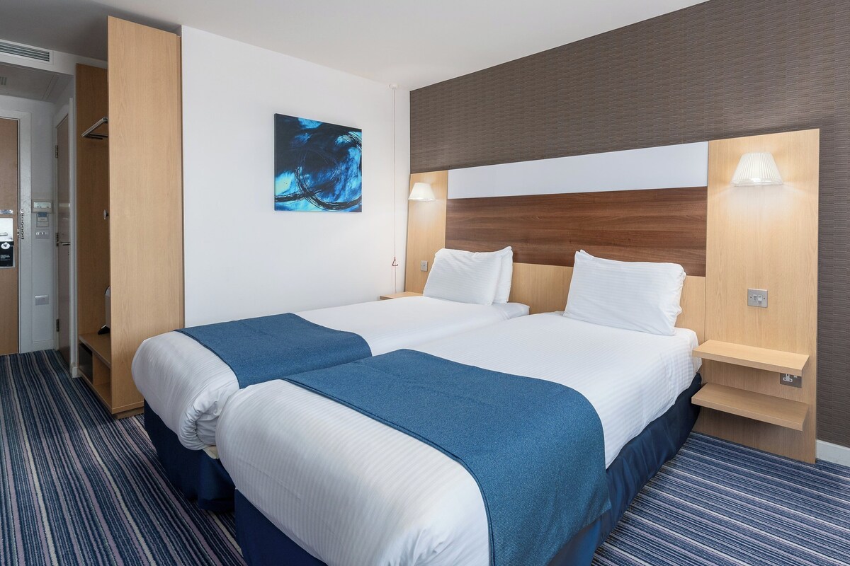 A hotel room with two beds, a large abstract painting, a wooden headboard, a nightstand, and a carpeted floor.