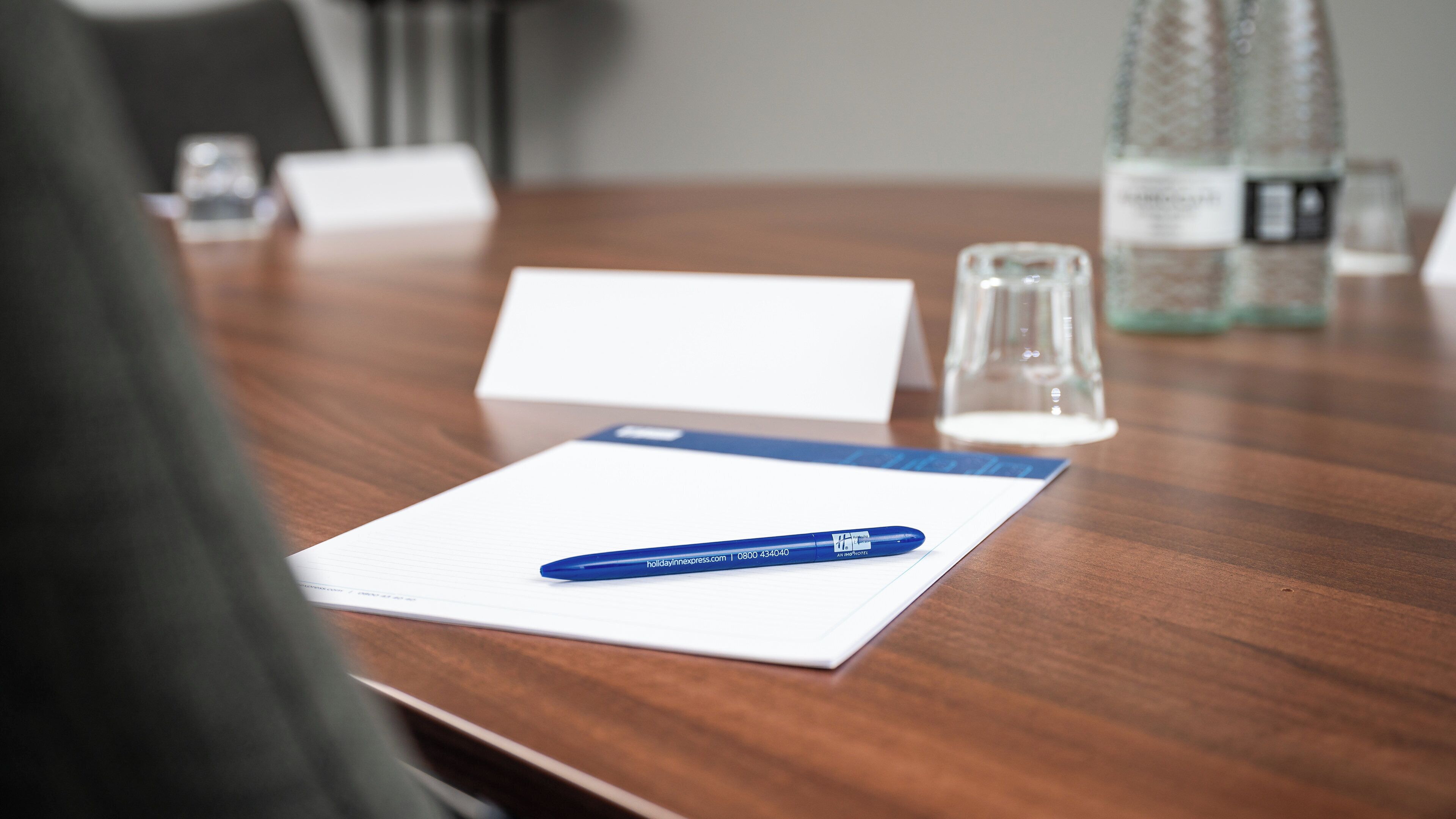 A wooden conference table with a notepad, a pen, a glass, and a bottle.