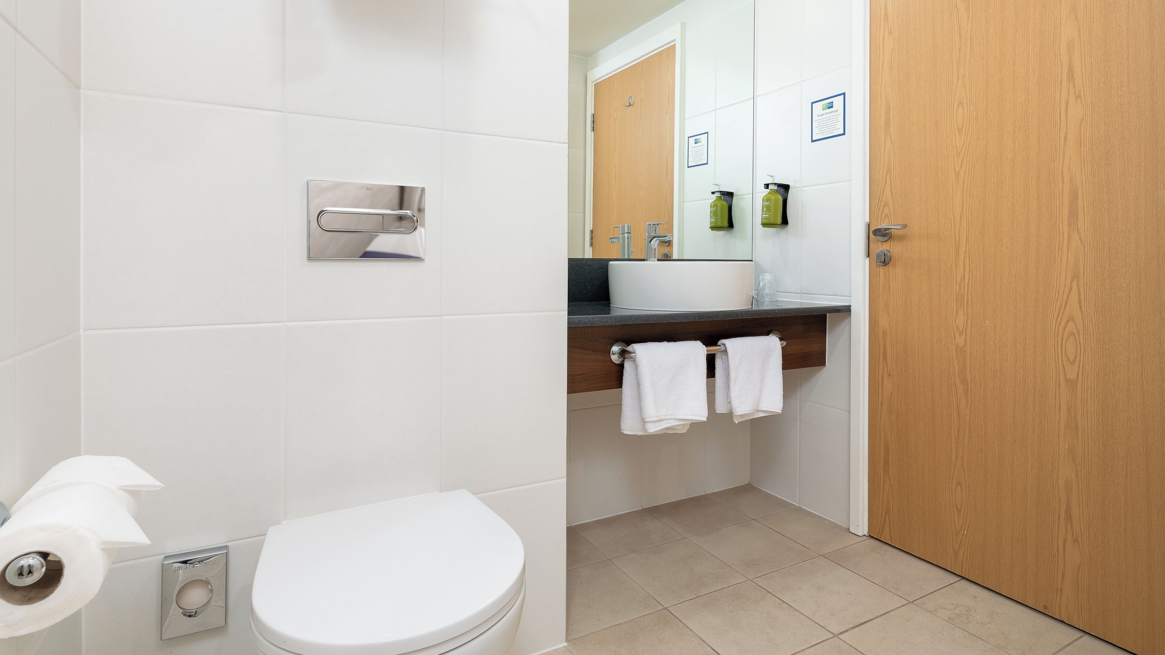 A bathroom with a toilet, a sink, and a mirror. There are towels hanging on a rack and a wooden door.