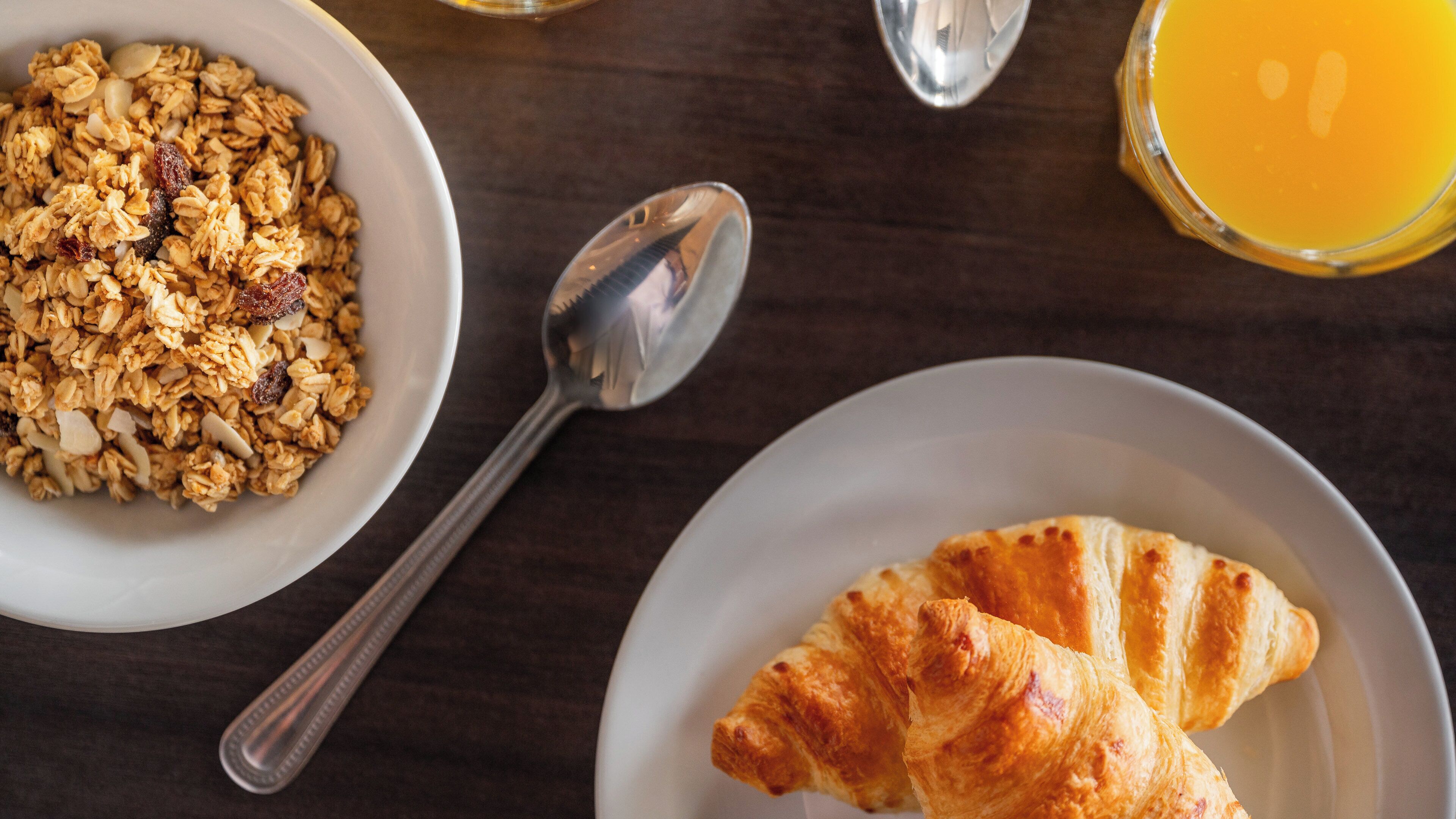 A breakfast setting with a bowl of granola, a croissant, a glass of orange juice, and a spoon on a wooden table.