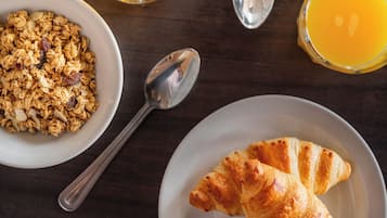 A breakfast setting with a bowl of granola, a croissant, a glass of orange juice, and a spoon on a wooden table.