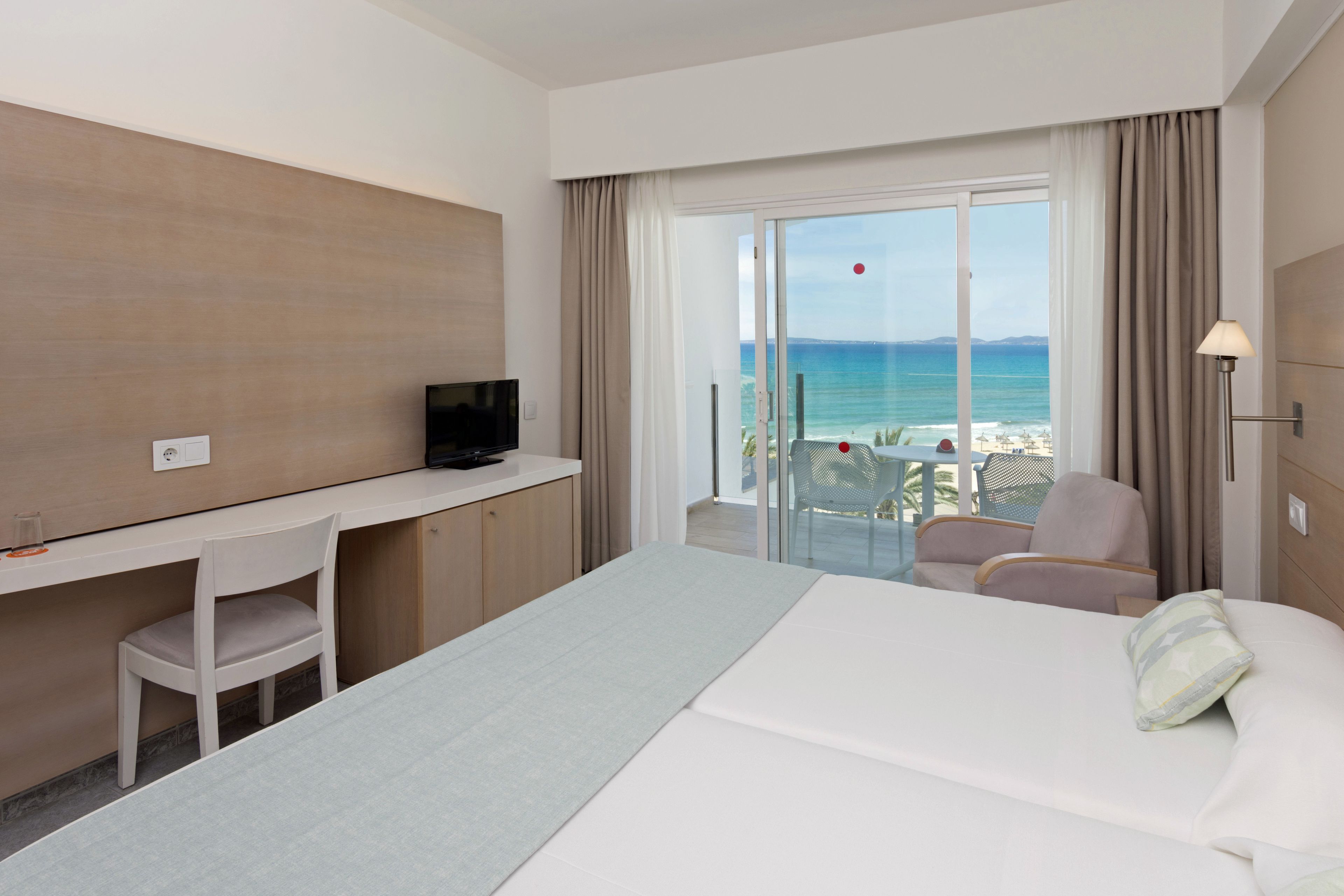 A hotel room with a bed, a desk, a chair, a TV, and a view of the beach.