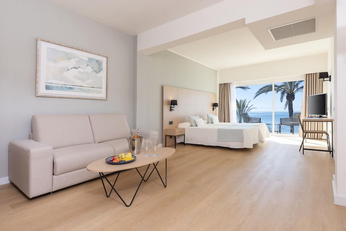 Een moderne hotelkamer met een bank, een eettafel met fruit en glazen, een bed met uitzicht op de oceaan en een televisie.