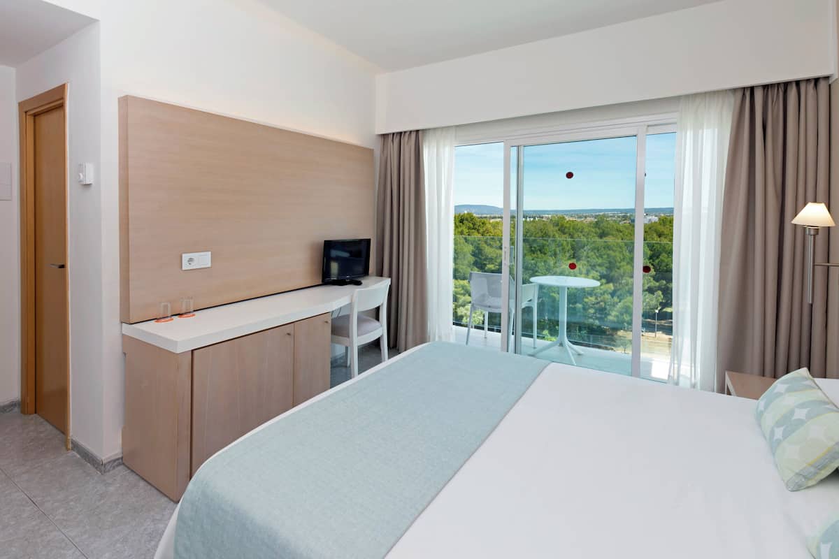Een hotelkamer met een bed, bureau, stoel, televisie en een balkon met uitzicht op groen.