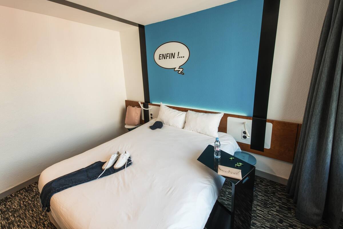 A hotel room with a bed, a nightstand, a wall with a blue accent, and a window with curtains.