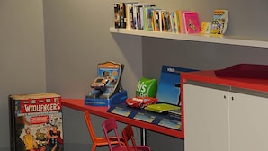 A children's play area with a red table, colorful chairs, a vending machine, and a bookshelf with various books.