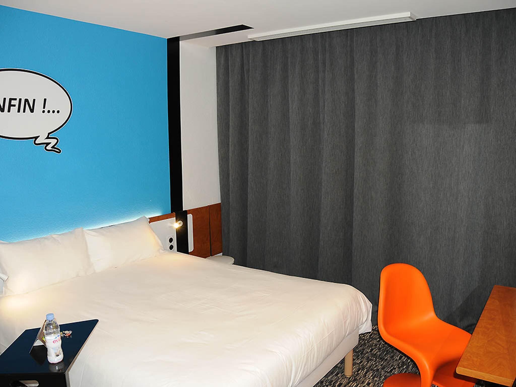 Una habitación de hotel con una cama, una mesita de noche, una botella de agua, una pared azul, una cortina gris y una silla naranja.