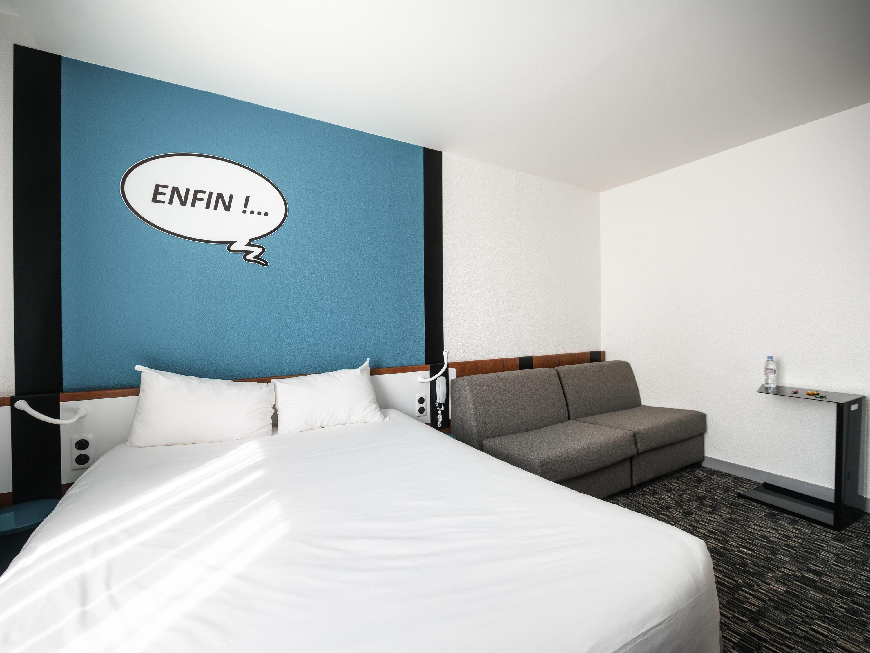 A hotel room with a bed, a sofa, a wall with a blue accent, and a speech bubble with text.