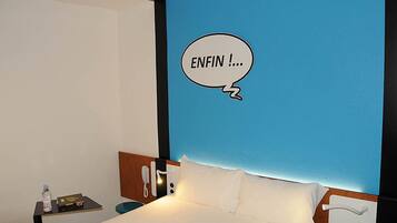 A hotel room with a bed, a bedside table with a bottle and a book, a phone, and a blue wall with a speech bubble saying 'ENFIN !'.