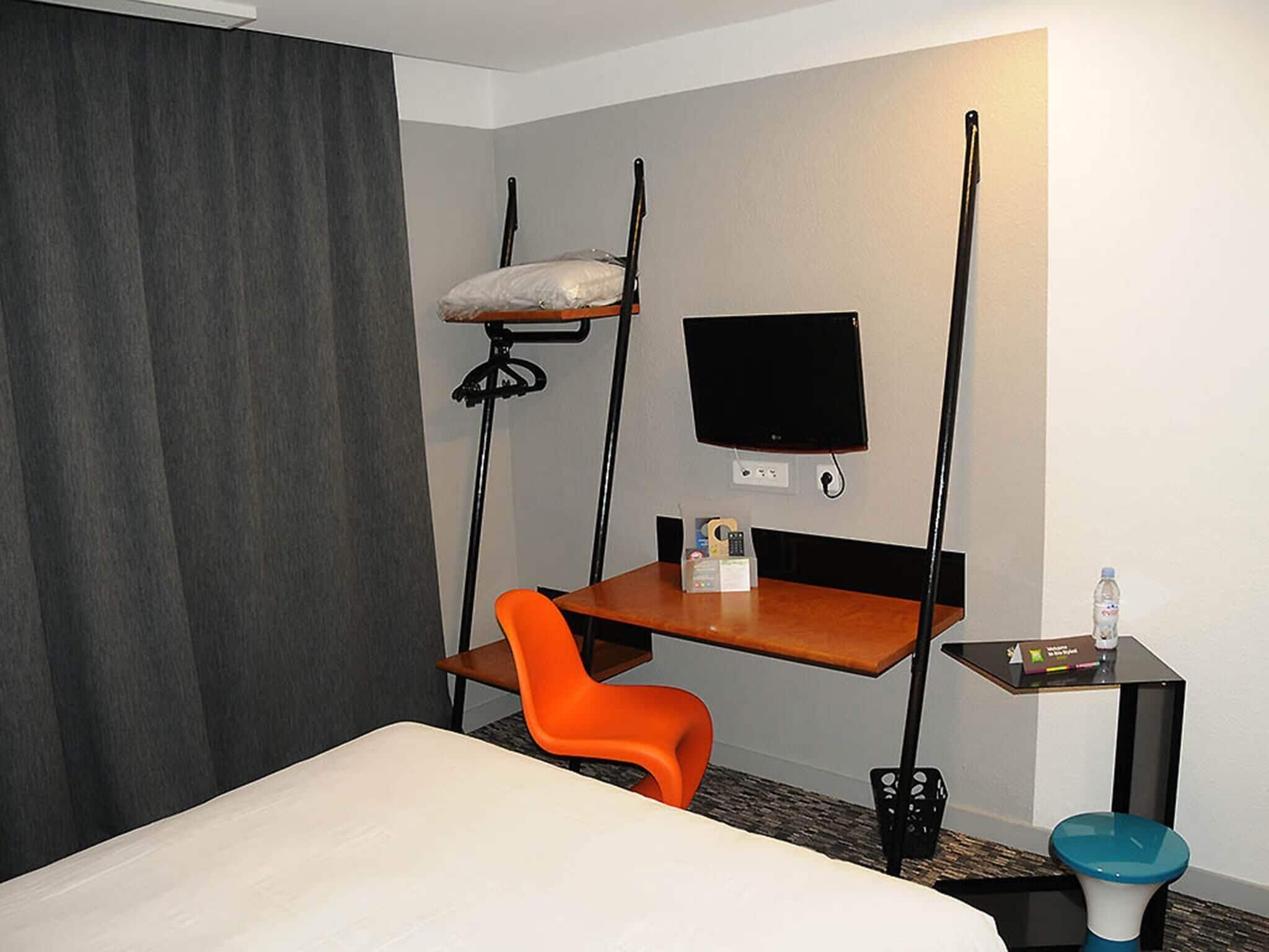 Una camera d'albergo con un letto, una scrivania, una poltrona arancione, una televisione, una bottiglia d'acqua e un cestino.