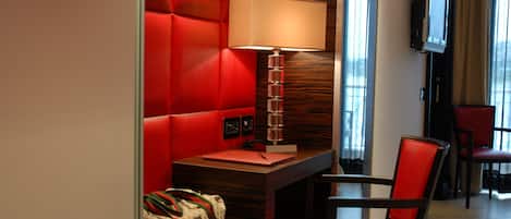 Une chambre d’hôtel avec une tête de lit rembourrée de tissu rouge, un bureau avec une lampe, un téléviseur fixé au mur et une chaise rouge.