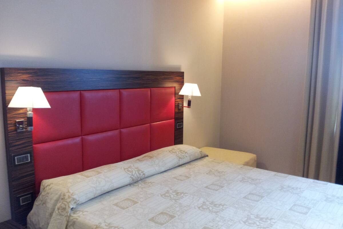 Una camera d'hotel con una testiera imbottita rossa, due lampade da comodino, un letto con copripiumino decorato e un tavolino di sostegno.
