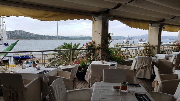 An outdoor dining area with tables and chairs, overlooking a body of water and hills. - Hotel Excelsior (La Maddalena)