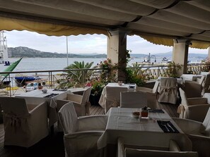 An outdoor dining area with tables and chairs, overlooking a body of water and hills. - Hotel Excelsior (La Maddalena)
