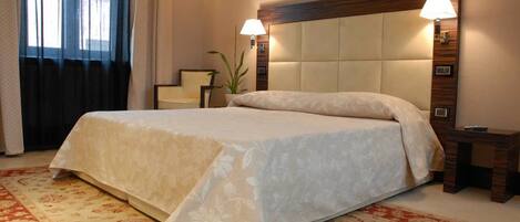 A hotel room with a large bed, a wooden headboard, two bedside lamps, a small table, and a potted plant.