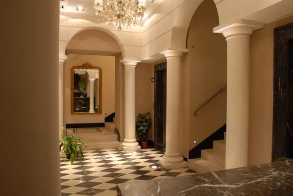 Un amplio pasillo con piso de cuadros, columnas blancas y una escalera que sube a un nivel superior.