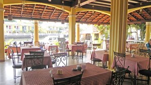 An outdoor restaurant with checkered tablecloths, metal chairs, and a view of buildings and palm trees. - Riverside Regency Resort (Baga)