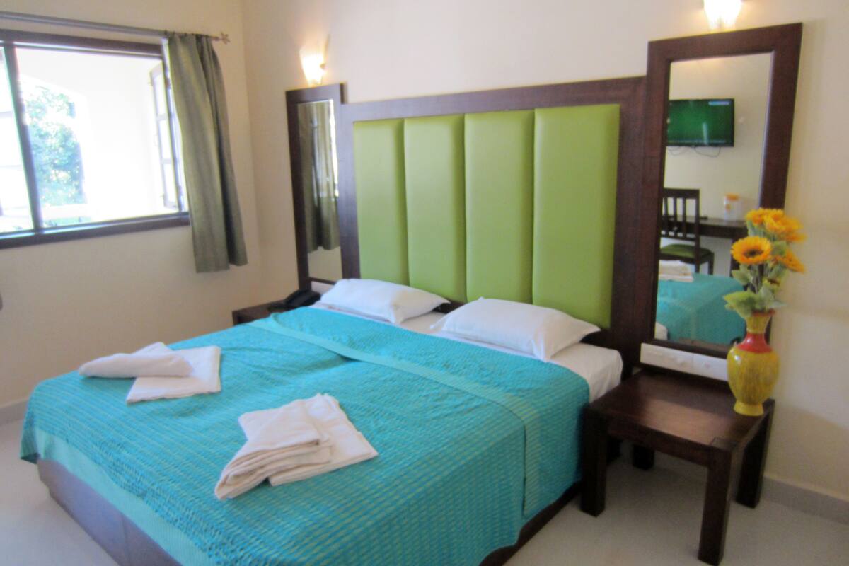 A neatly made bed with a green headboard, a bedside table with a vase of flowers, and a large mirror on the wall.