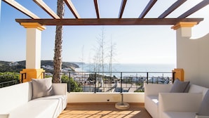 A balcony with a white sofa, a round table, and a view of the sea and hills. - Apartamentos Turísticos The View (Vila do Bispo)