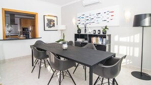 A modern dining area with a long table, six chairs, a potted plant, a bookshelf, and a wall-mounted artwork. - Apartamentos Turísticos The View (Vila do Bispo)