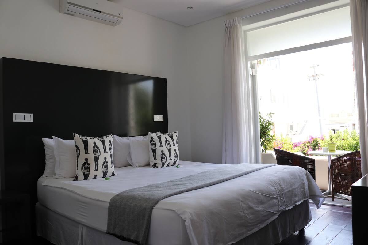 A hotel room with a large bed, white bedding, a black headboard, and a view of a balcony with plants and a table.