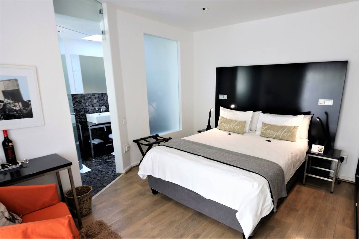 A modern bedroom with a large bed, a bedside table, a chair, and a small table with a bottle.