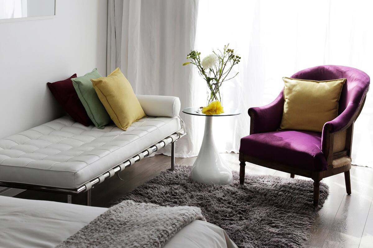 A modern bedroom with a bed, a sofa, a purple armchair, a small table with a vase, and a large window with sheer curtains.