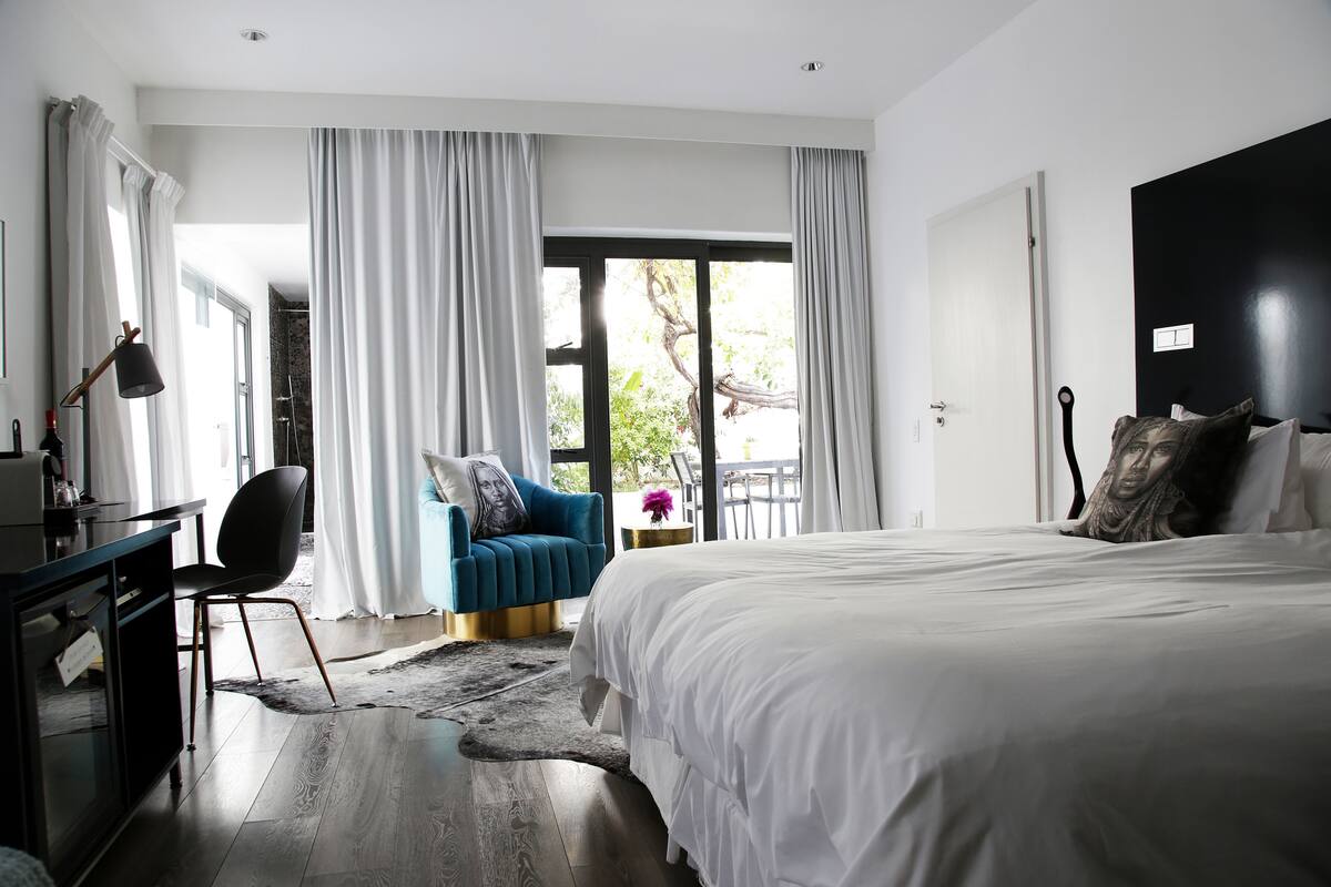 A modern hotel room with a large bed, a desk, and a chair. There is a view of greenery outside the window.