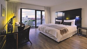 Quarto de hotel moderno com uma cama grande, uma mesa de trabalho com luminária e vista para a cidade através de amplas janelas.