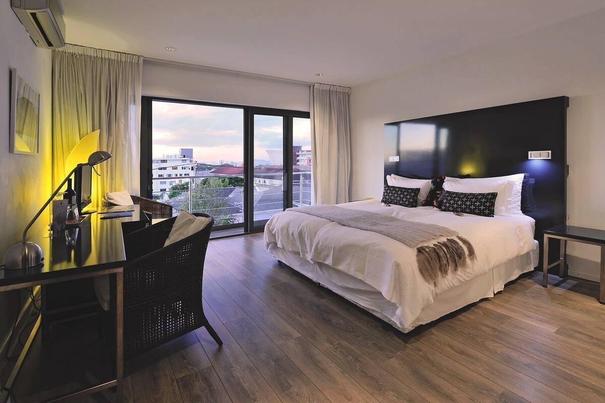 A modern hotel room with a large bed, a desk with a lamp, and a view of the city through large windows.