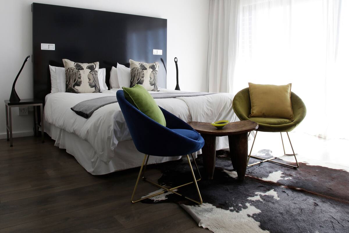 A modern hotel room with a large bed, a blue armchair, a green armchair, a small round table, and a black and white rug.