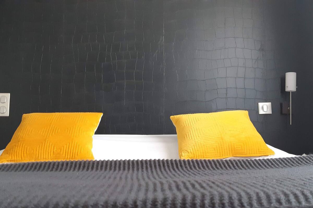 A hotel room with a black textured wall, two yellow pillows, a white bedspread, and a grey blanket.