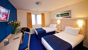 Une chambre d’hôtel avec deux lits, un bureau, une chaise, un vase avec des fleurs, un tableau au mur et une fenêtre avec des rideaux.