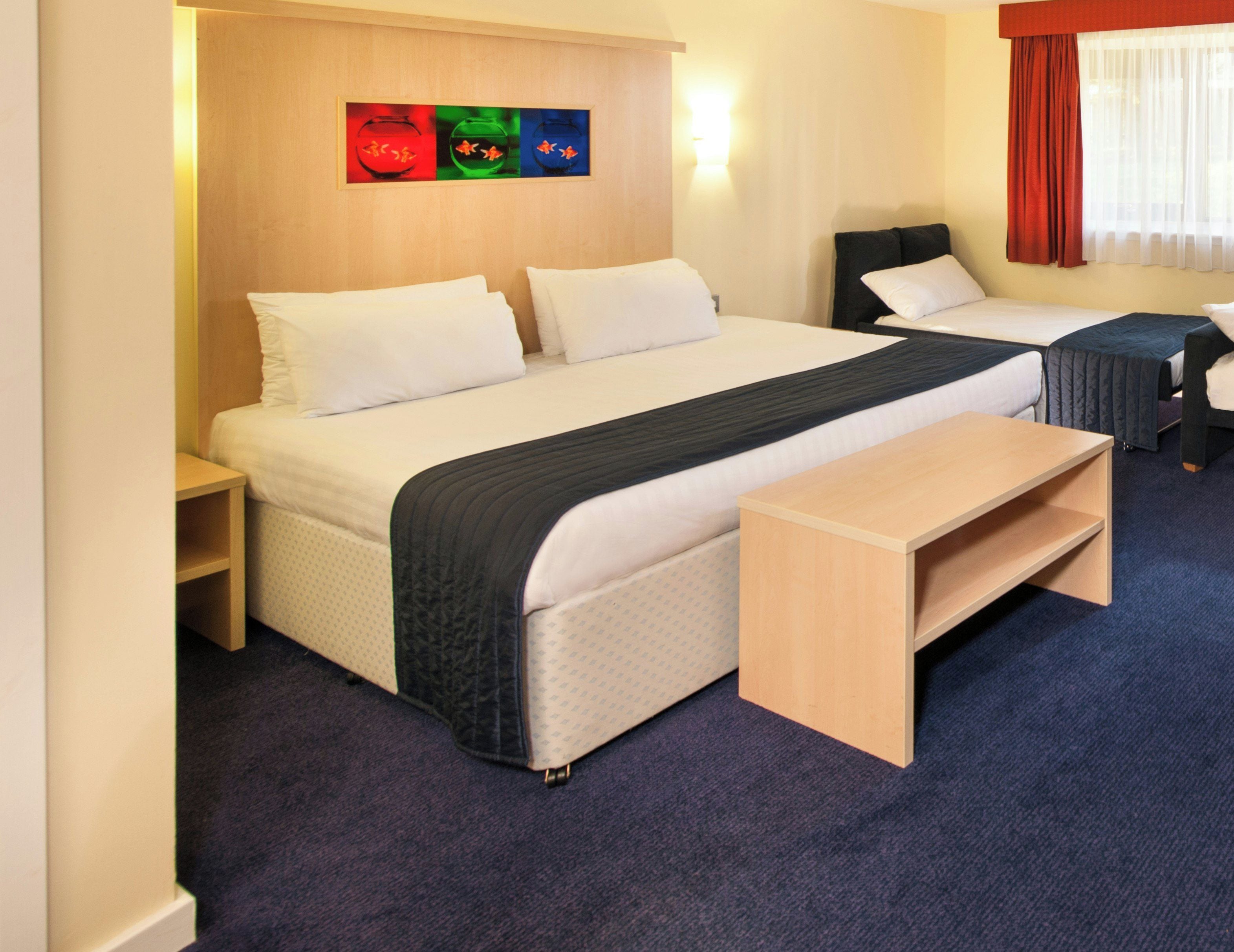 A hotel room with a large bed, a bedside table, a bench, and a wall-mounted artwork.