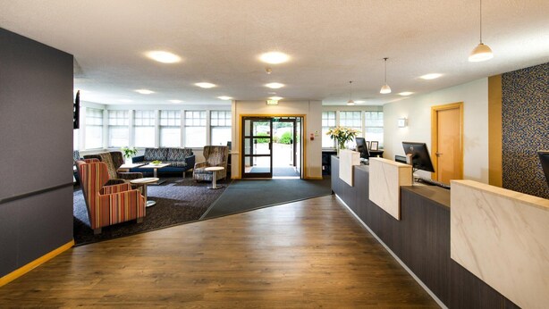 A reception area with a wooden floor, a reception desk, a sofa, and a patterned wall. - Leonardo Inn Aberdeen Airport (Aberdeen)