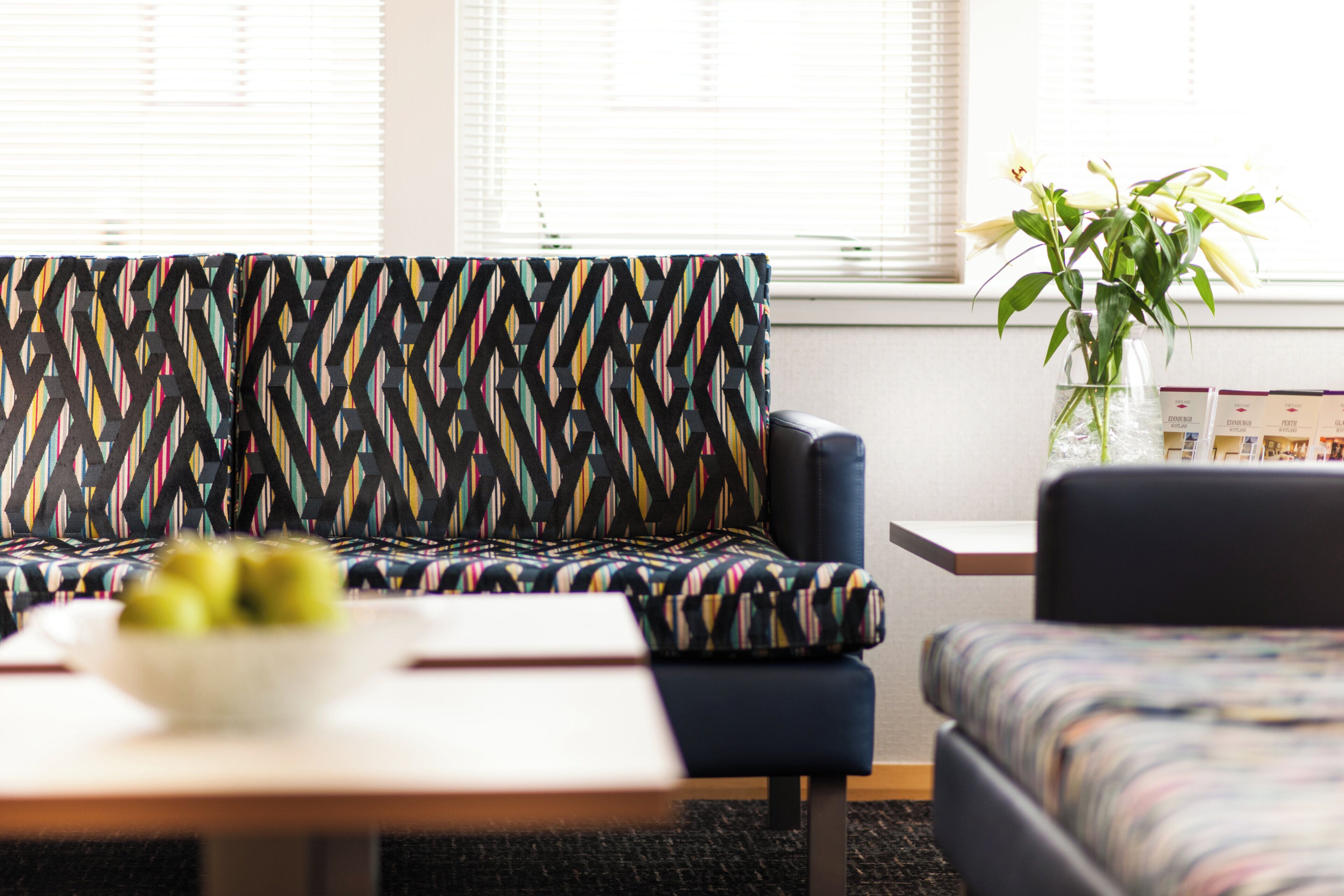 A modern living room with a patterned sofa, a small table with a bowl of fruit, and a vase with flowers.