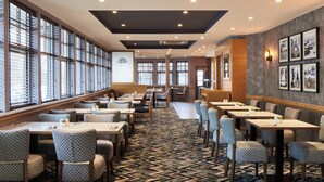 Ein Hotel-Speisesaal mit mehreren Tischen und Stühlen, großen Fenstern und einem gemusterten Teppich.