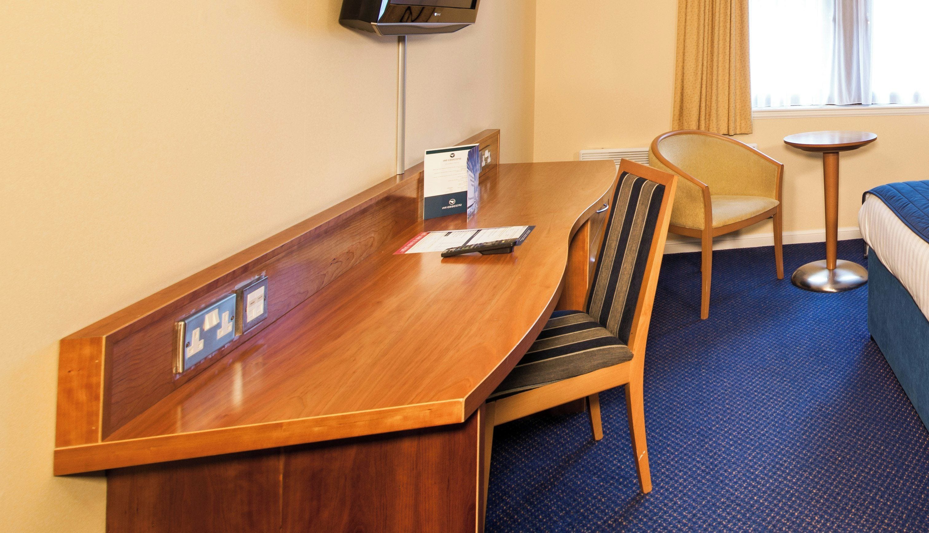 A hotel room with a wooden desk, a chair, a bed, a small round table, and a television mounted on the wall.