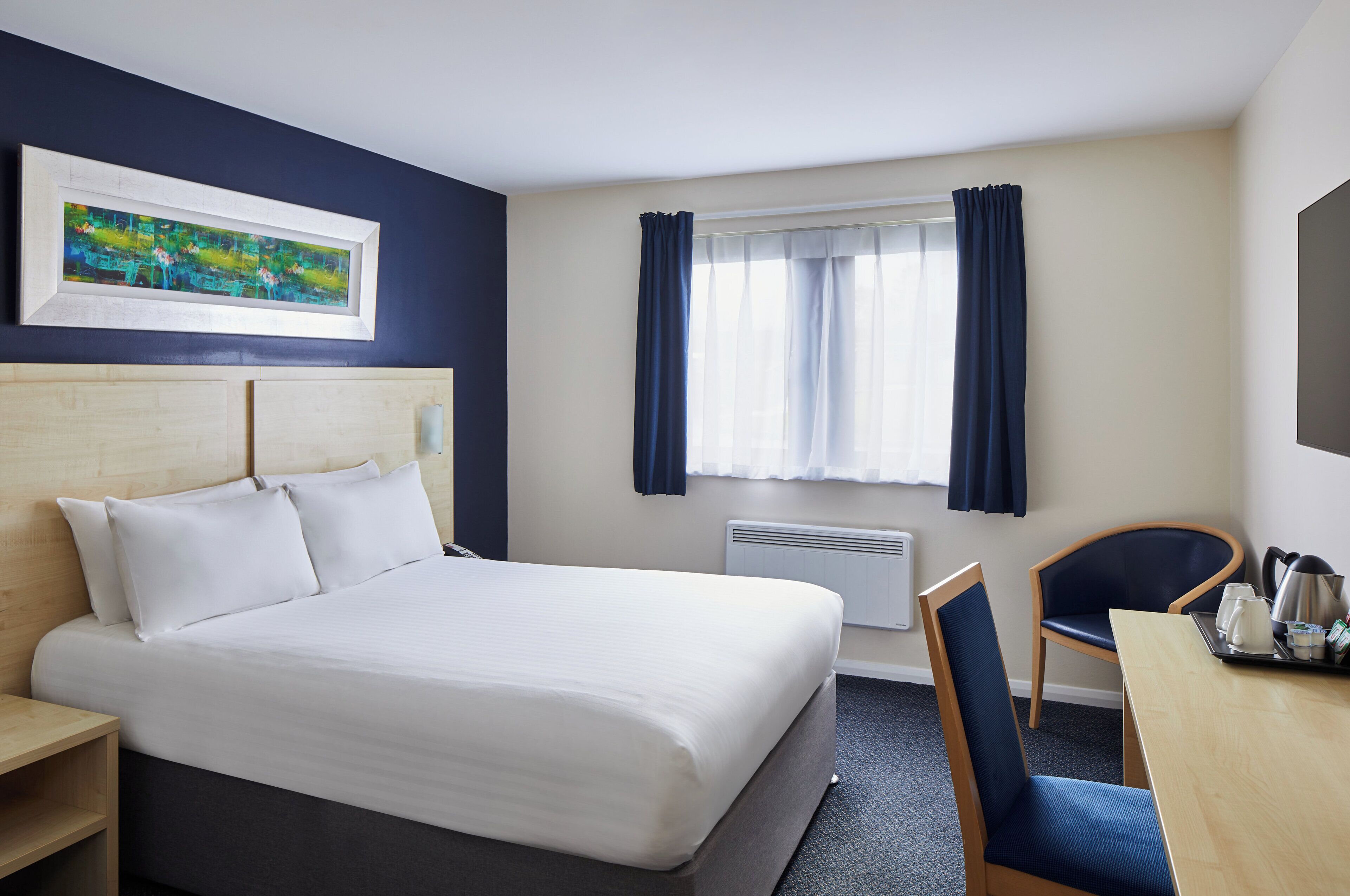 Ein Hotelzimmer mit Bett, Schreibtisch, Stuhl, einem Bild an der Wand und einem Vorhangfenster.