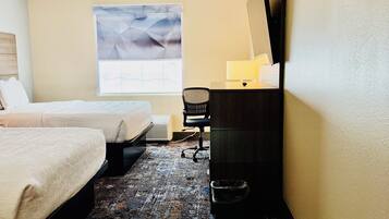 Una camera d'albergo con due letti, una scrivania con una lampada, una sedia, un'ampia finestra con tenda geometrica e pavimento con moquette.