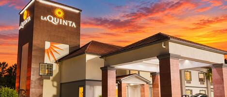 A hotel entrance with a prominent sign, covered parking, and a clear sky.