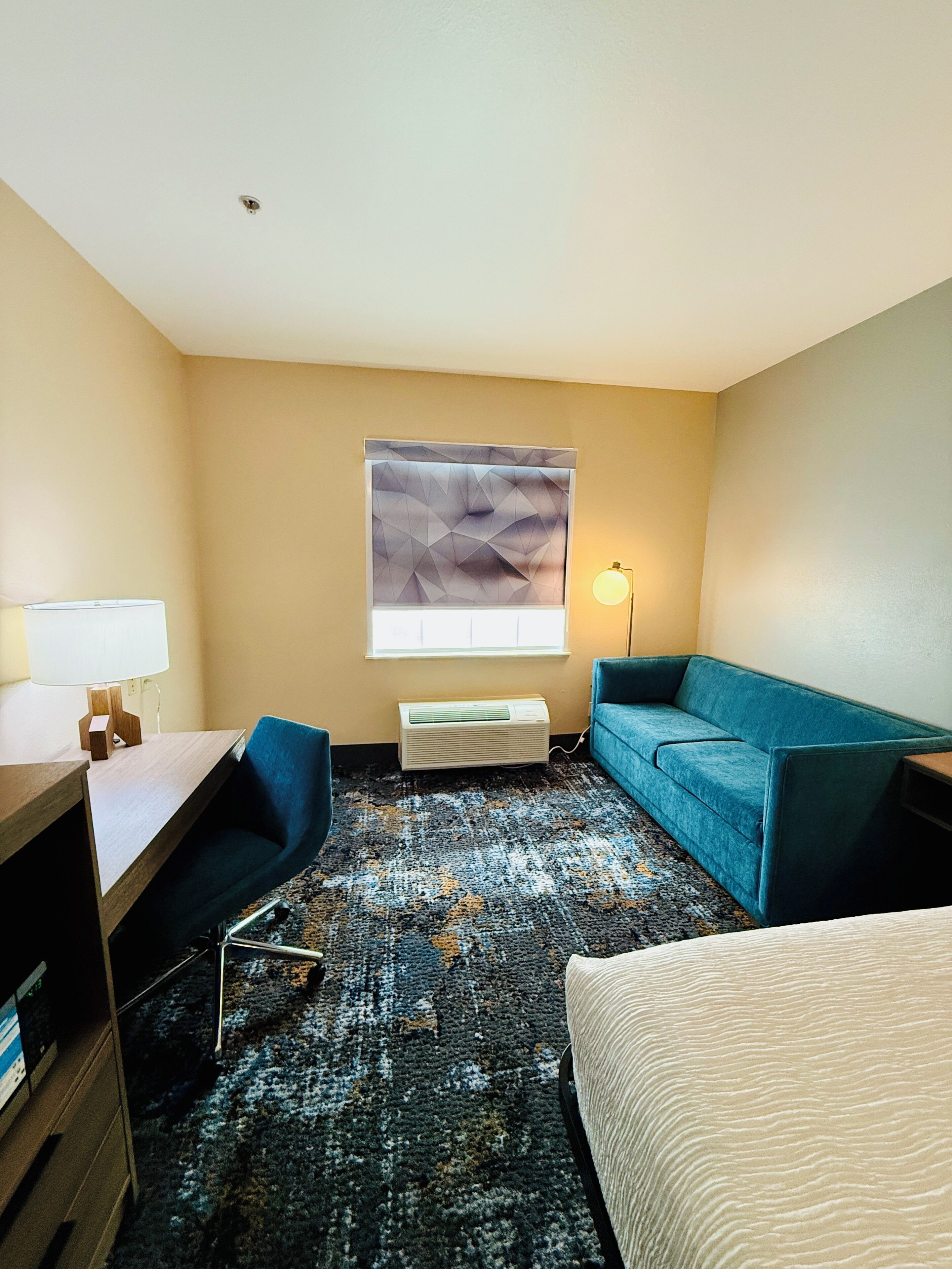 A hotel room with a desk, a blue sofa, a large window with blinds, a standing lamp, and a patterned carpet.